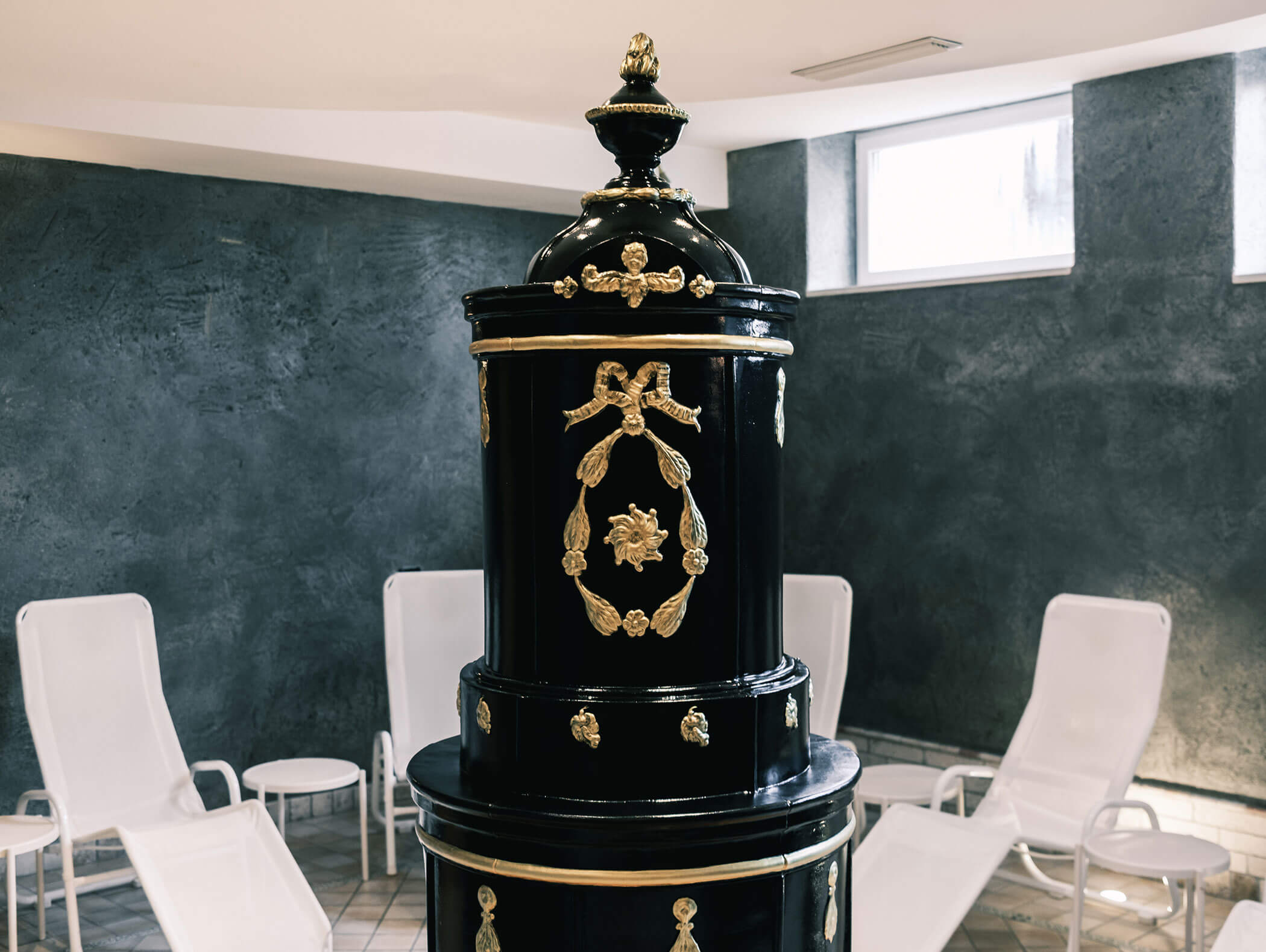 In einem Raum mit weißen Sesseln und grauen Wänden steht ein schwarz-goldener, kunstvoll verzierter Ofen. - Hotel Kristiania