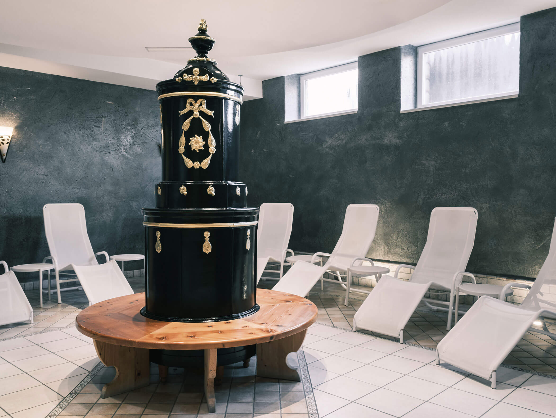 In der Mitte steht ein schwarzer, kunstvoll verzierter Saunaofen, umgeben von weißen Liegestühlen in einem gefliesten, sanft beleuchteten Raum. - Hotel Kristiania