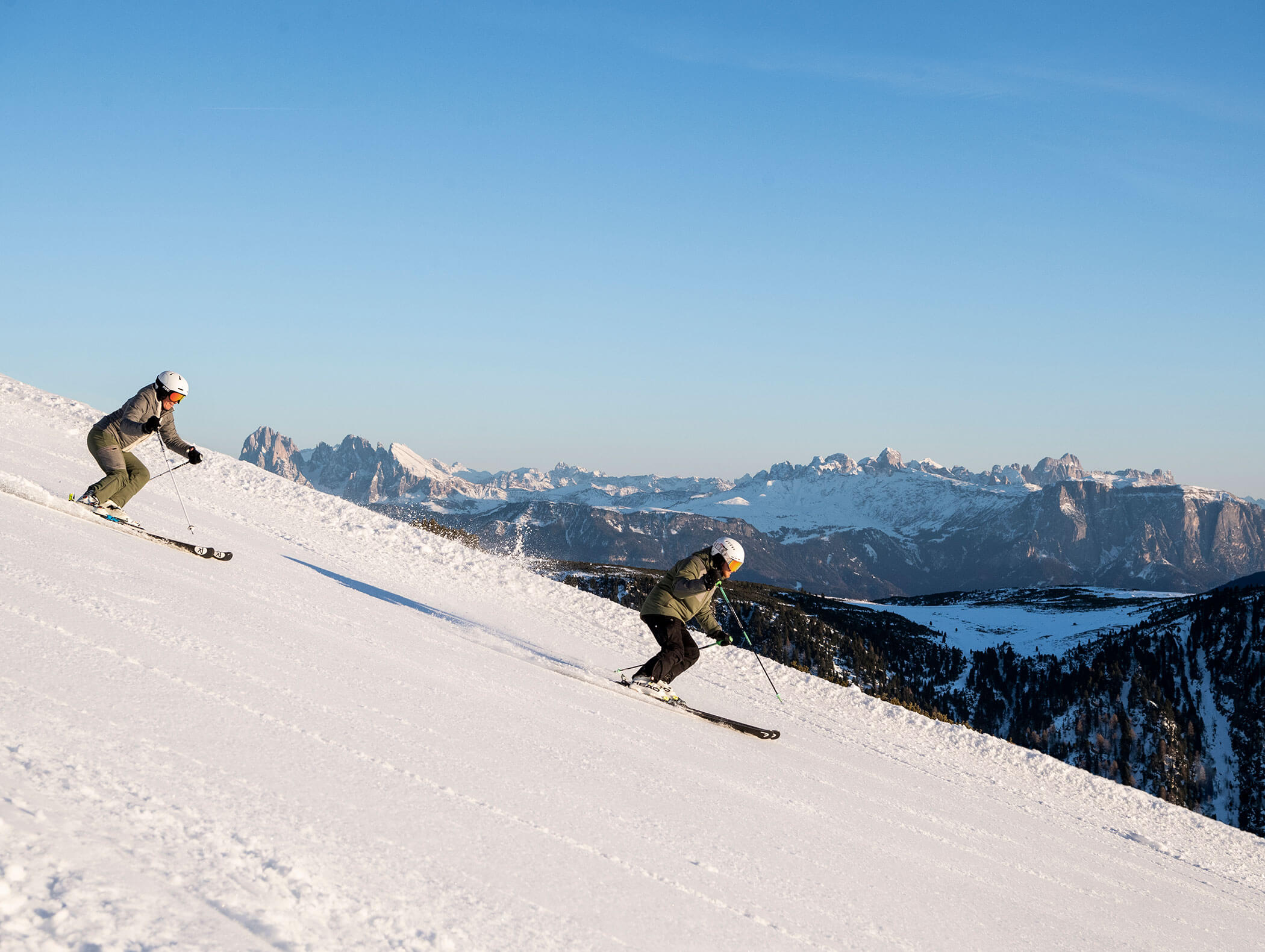 Due persone che scendono con gli sci lungo un pendio innevato, con le montagne in lontananza sotto un cielo azzurro e limpido. - Hotel Kristiania