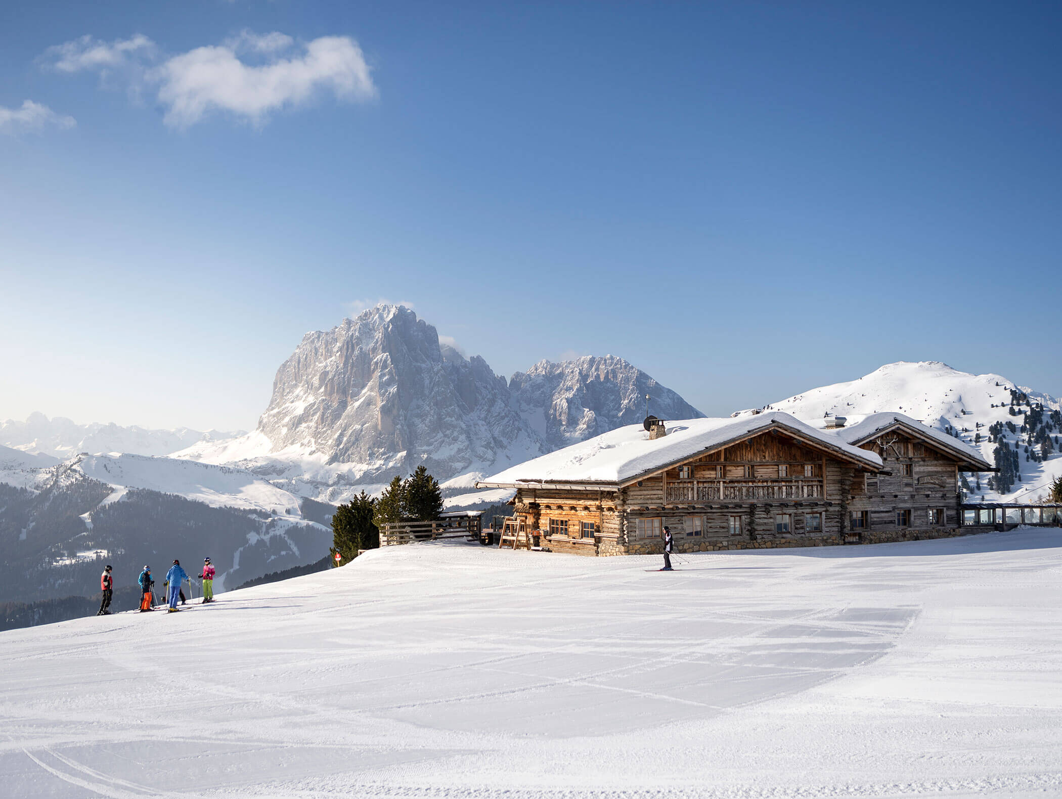 Sciatori nei pressi di un rifugio in legno su una montagna innevata, con una grande vetta rocciosa sullo sfondo. - Hotel Kristiania