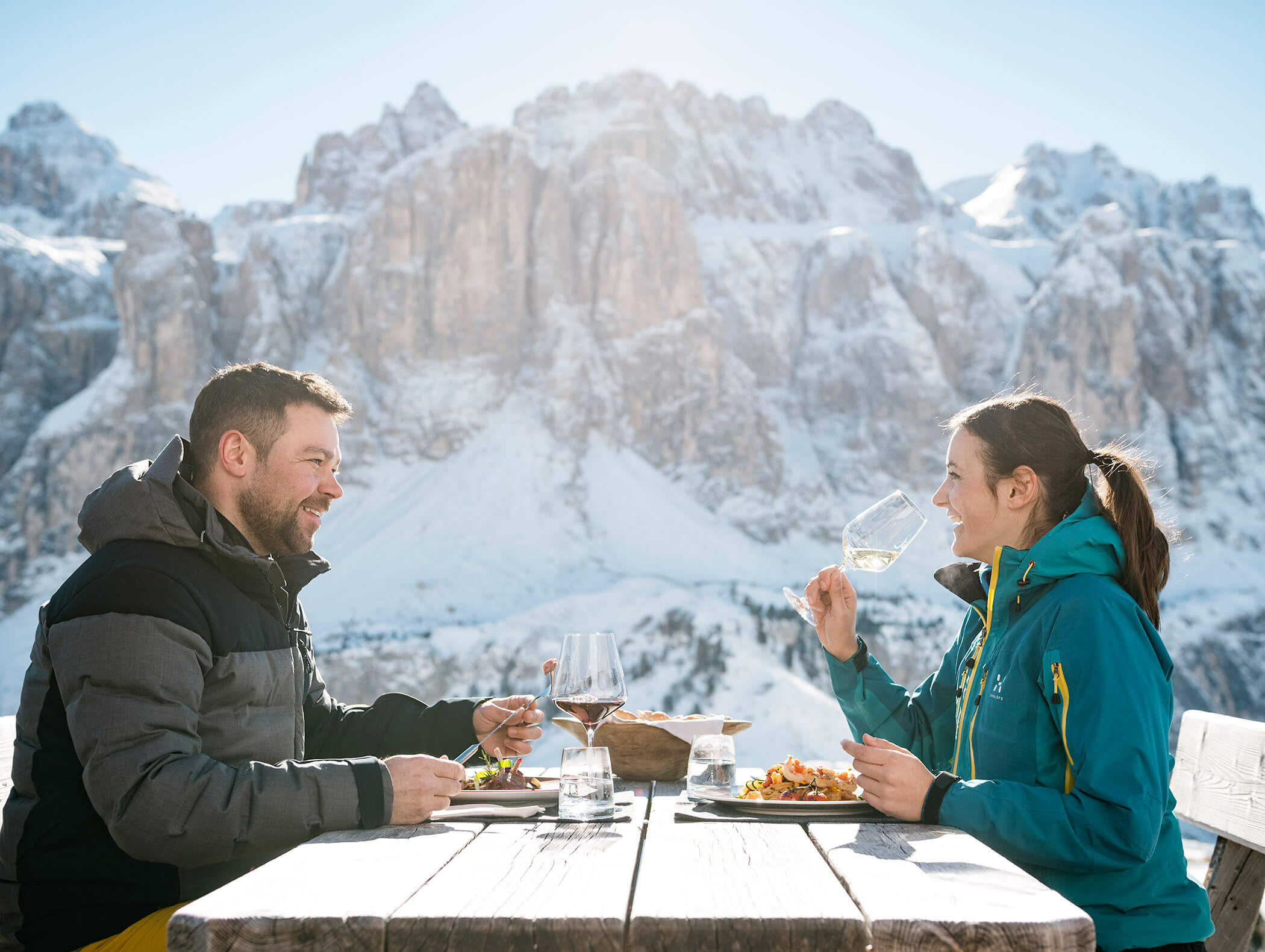 Un uomo e una donna si godono il cibo e il vino seduti a un tavolo all’aperto, con le montagne innevate sullo sfondo. - Hotel Kristiania