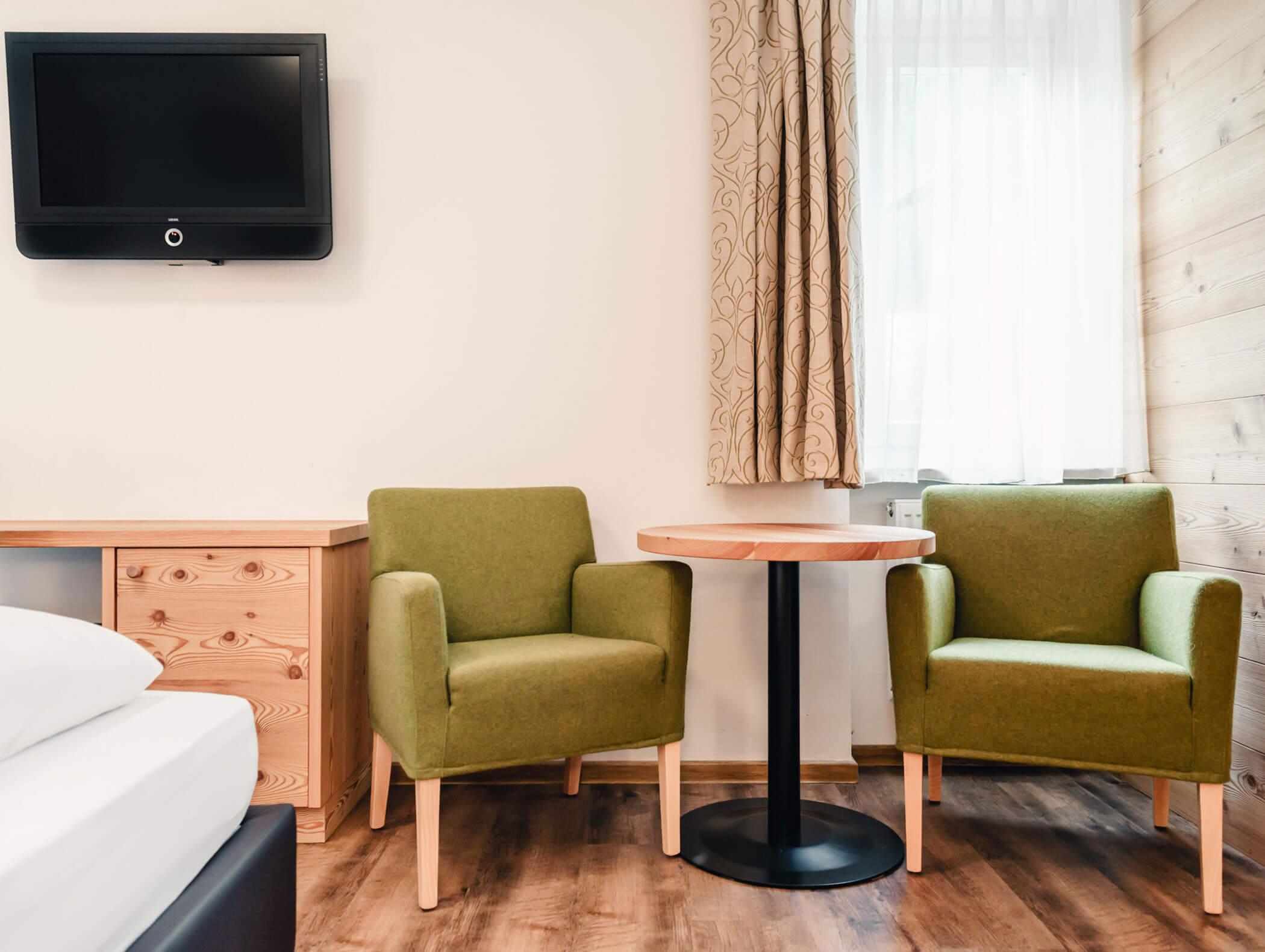 Due poltrone verdi e un tavolo rotondo vicino alla finestra in una stanza accogliente con una TV appesa alla parete. - Hotel Kristiania